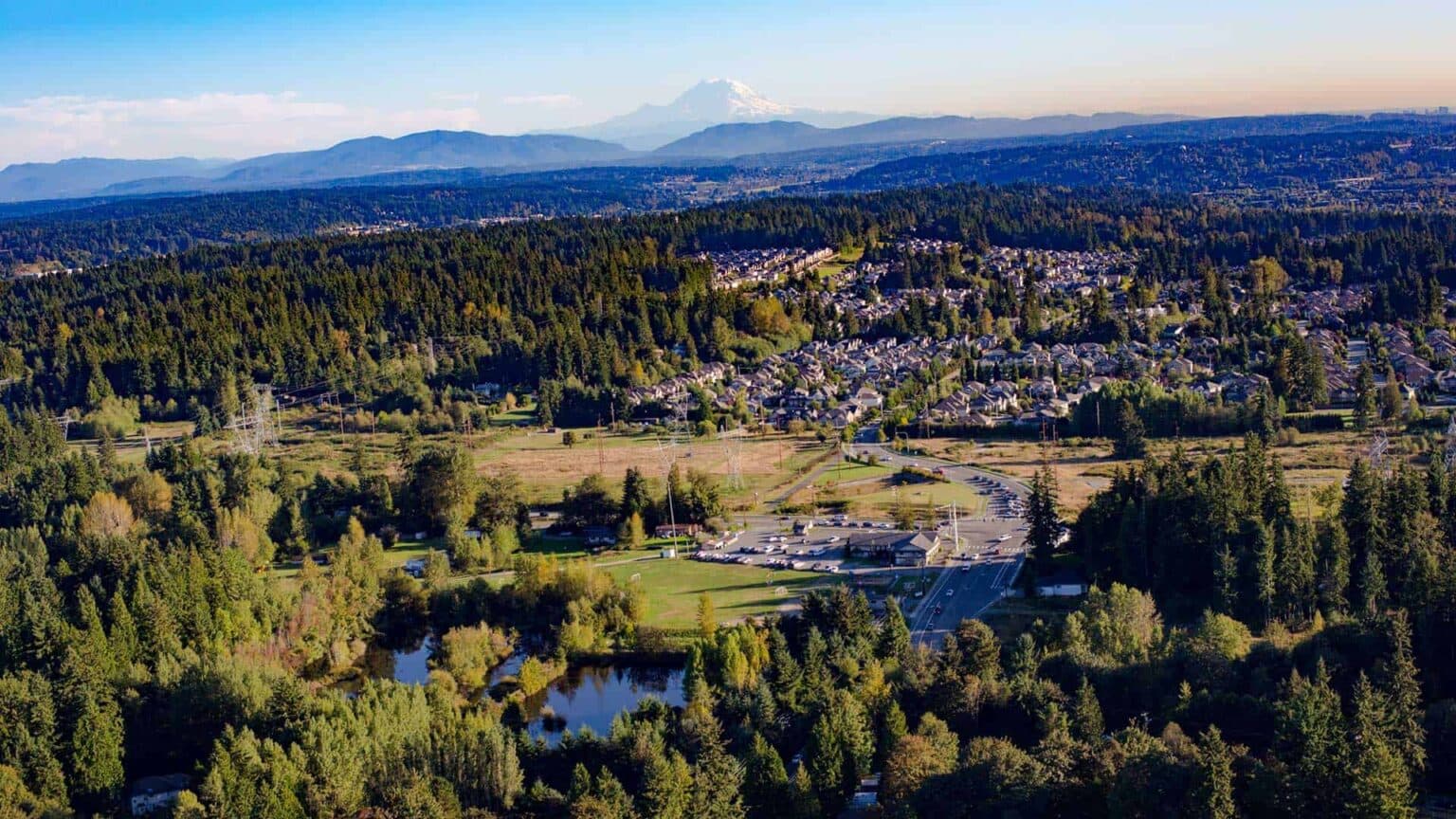 Aerial view of Bothell, Washington, showing a mix of forested areas, suburban neighborhoods, and roads with Mount Rainier visible in the background under a clear blue sky.