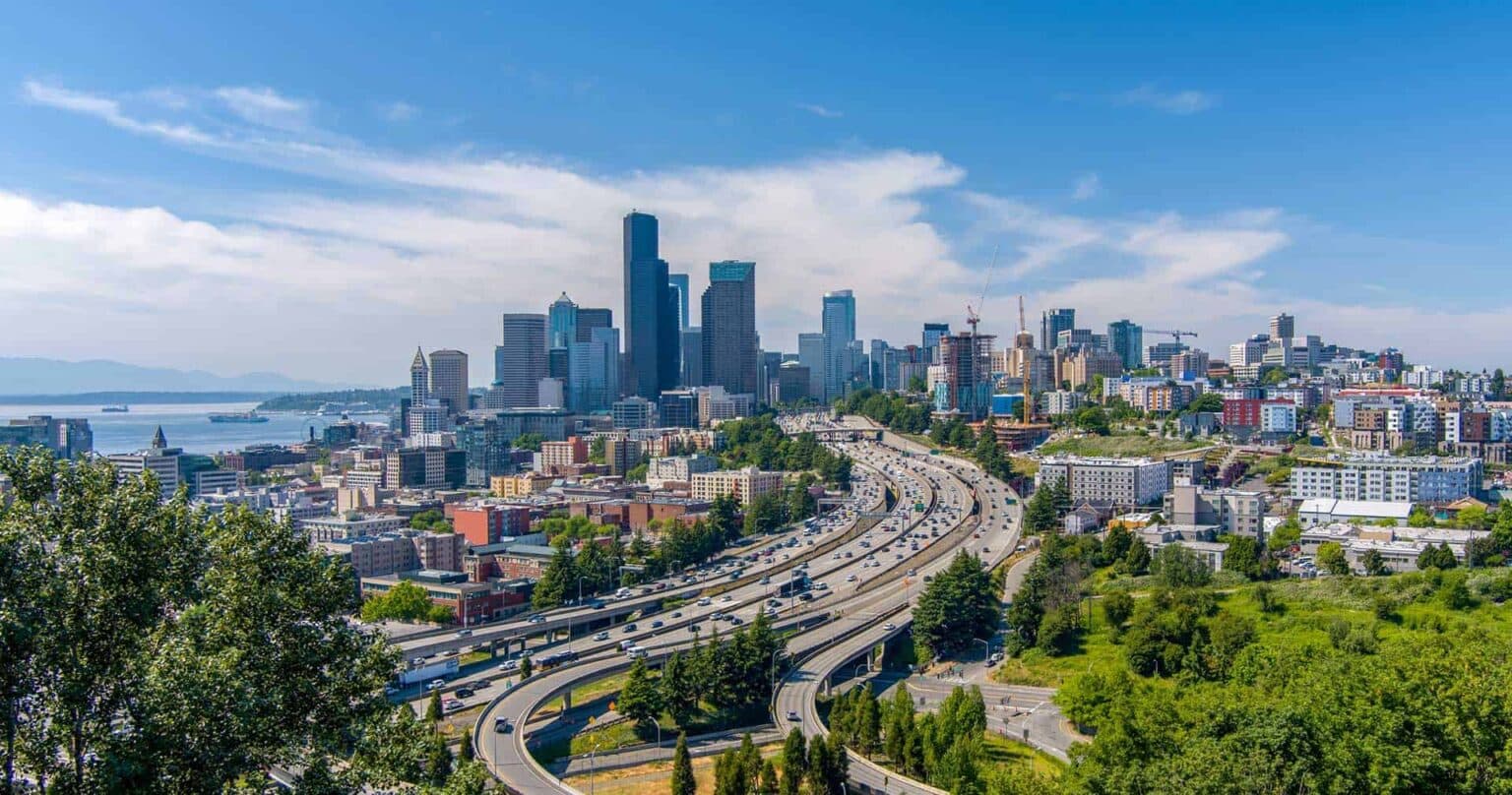 Aerial view of Seattle, Washington, showing a mix of forested areas, suburban neighborhoods, and roads with Mount Rainier visible in the background under a clear blue sky.